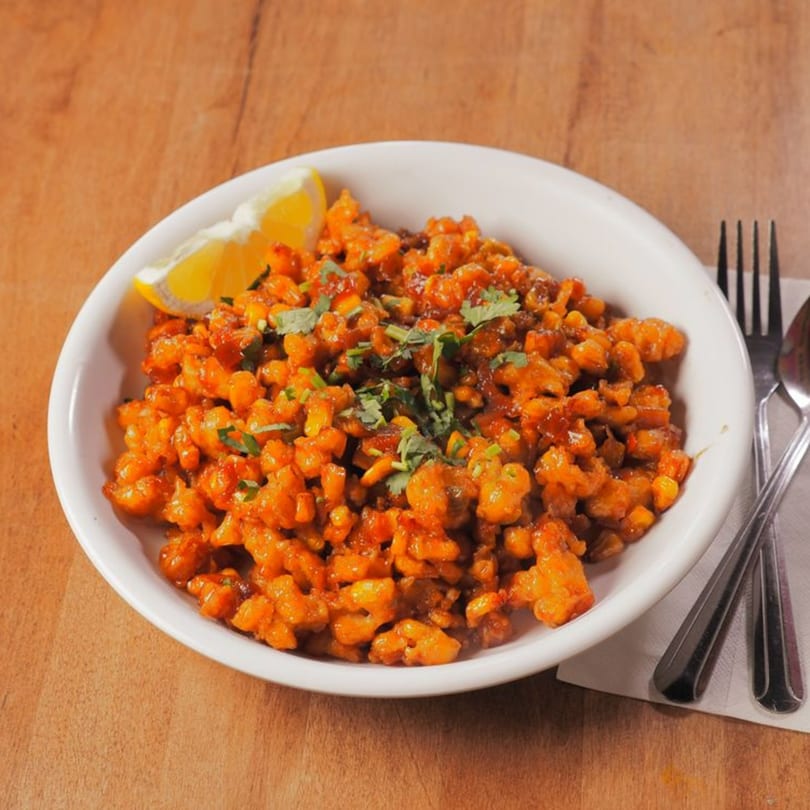 A white bowl of Pataka Indian Fusion Cuisine's spicy corn, garnished with cilantro and a lemon wedge.